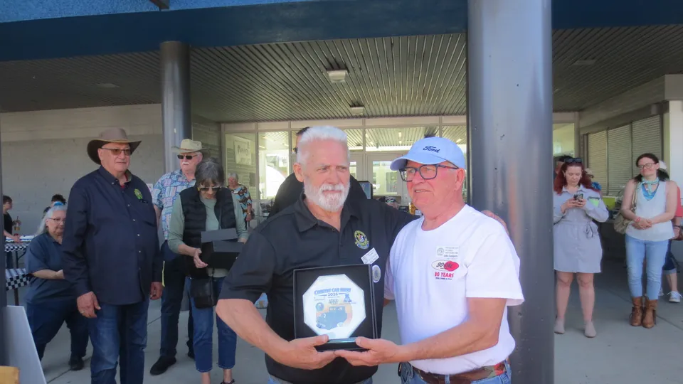 Fraser Field, in a black Country Car Show polo, holds the octagonal Country Car Show 2026 plaque beside a presenter in a Ford ball cap and white "90 Years" shirt at the Aldergrove School entrance, with onlookers behind.
