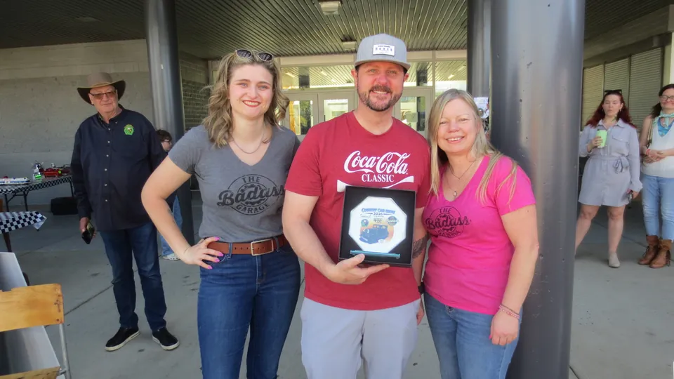 Ryan Schram, in a red Coca-Cola shirt, holds the octagonal Country Car Show 2026 plaque, flanked by two women in The Badass Garage shirts, outdoors at the show.