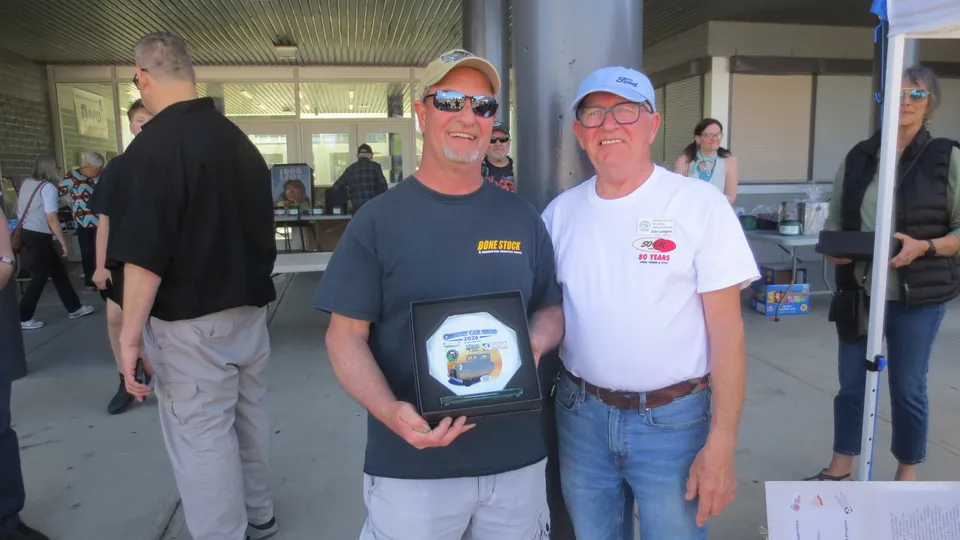 Terry Callewaert, in sunglasses, a ball cap and a dark "Bone Stock" t-shirt, holds the octagonal Country Car Show 2026 plaque beside a presenter in a Ford ball cap and white "90 Years" shirt, with the show crowd and an event tent behind.